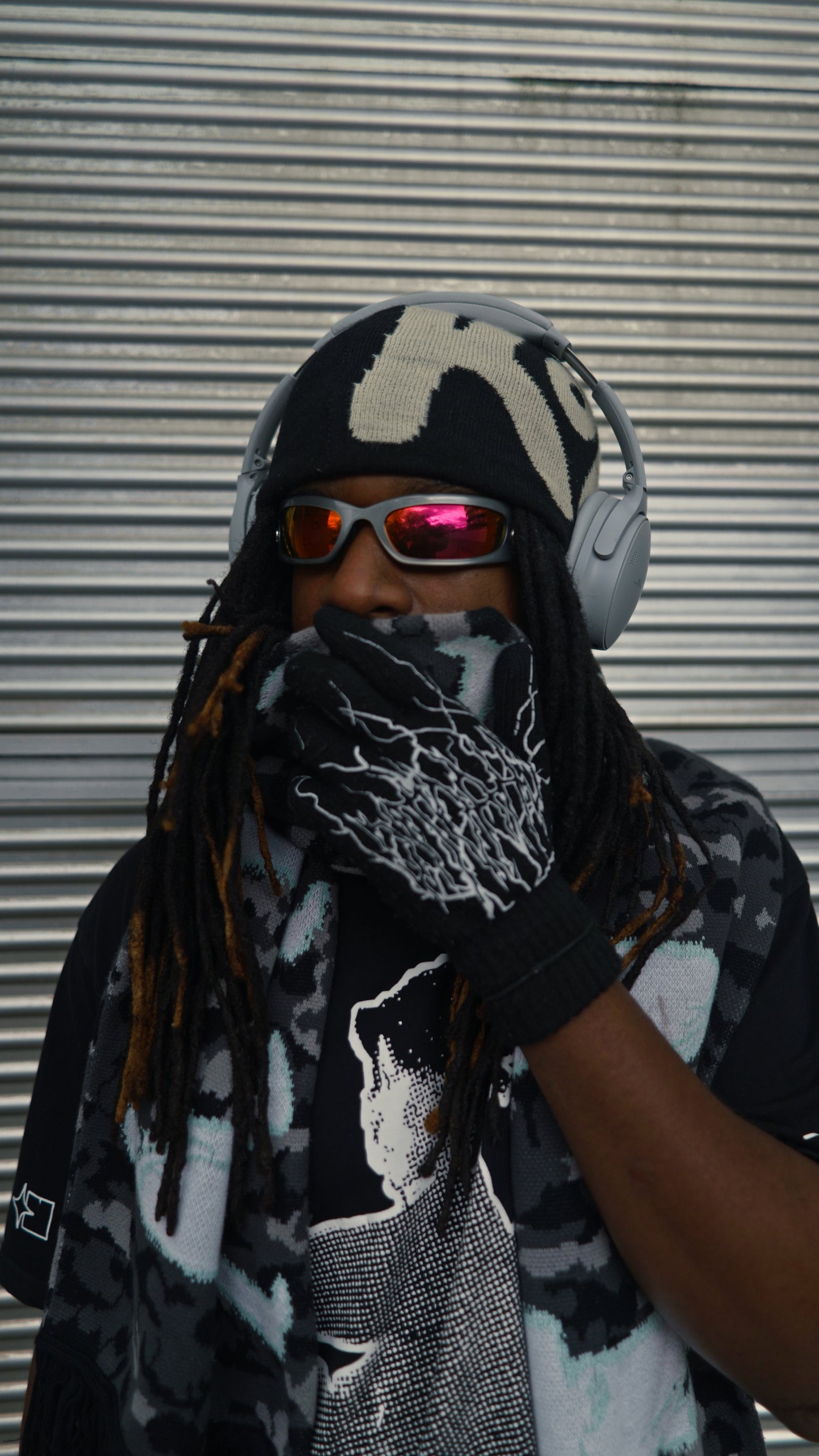 Person wearing a patterned KOZI beanie, sunglasses, and a scarf against a corrugated metal background